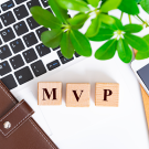 Blocos de madeira com as letras "MVP" sobre um teclado de notebook, simbolizando o conceito de Minimum Viable Product (Produto Mínimo Viável). Ao redor, elementos como uma carteira de couro, um smartphone e folhas verdes remetem à inovação e desenvolvimento de negócios.