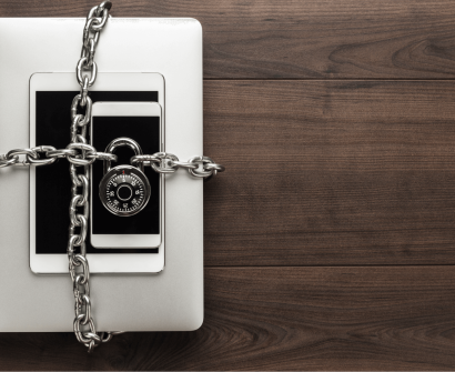 Dispositivos eletrônicos — laptop, tablet e smartphone — empilhados e envoltos por uma corrente de metal com cadeado, simbolizando a proteção de software e a segurança da propriedade intelectual.